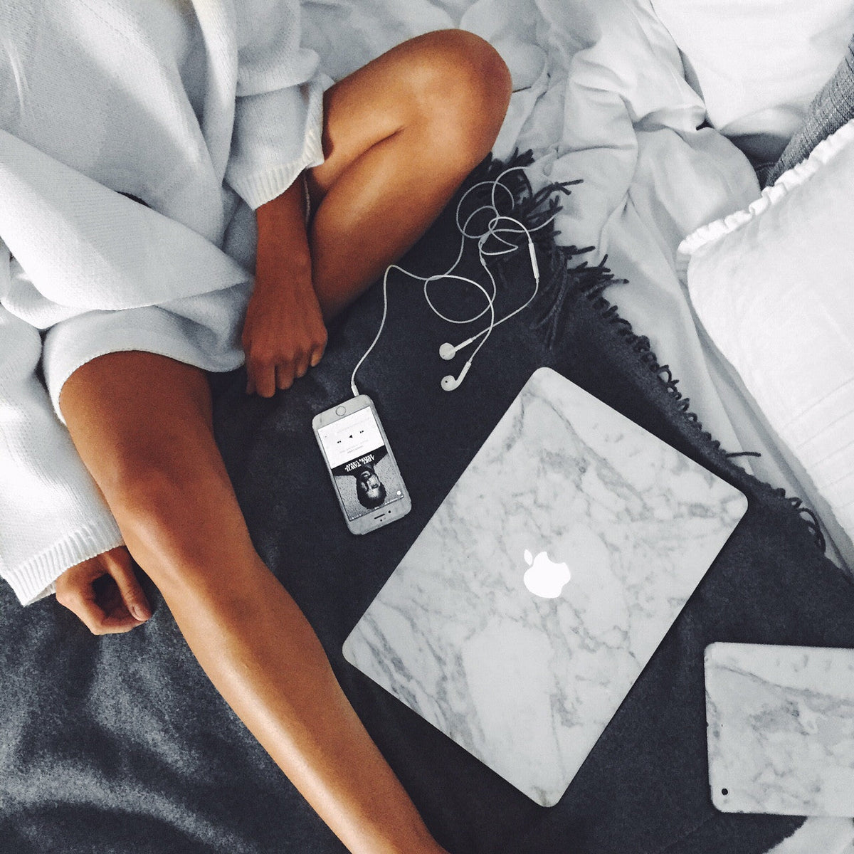 Person sitting on a bed with a white marble MacBook skin, smartphone, and wired earphones on a grey blanket.
