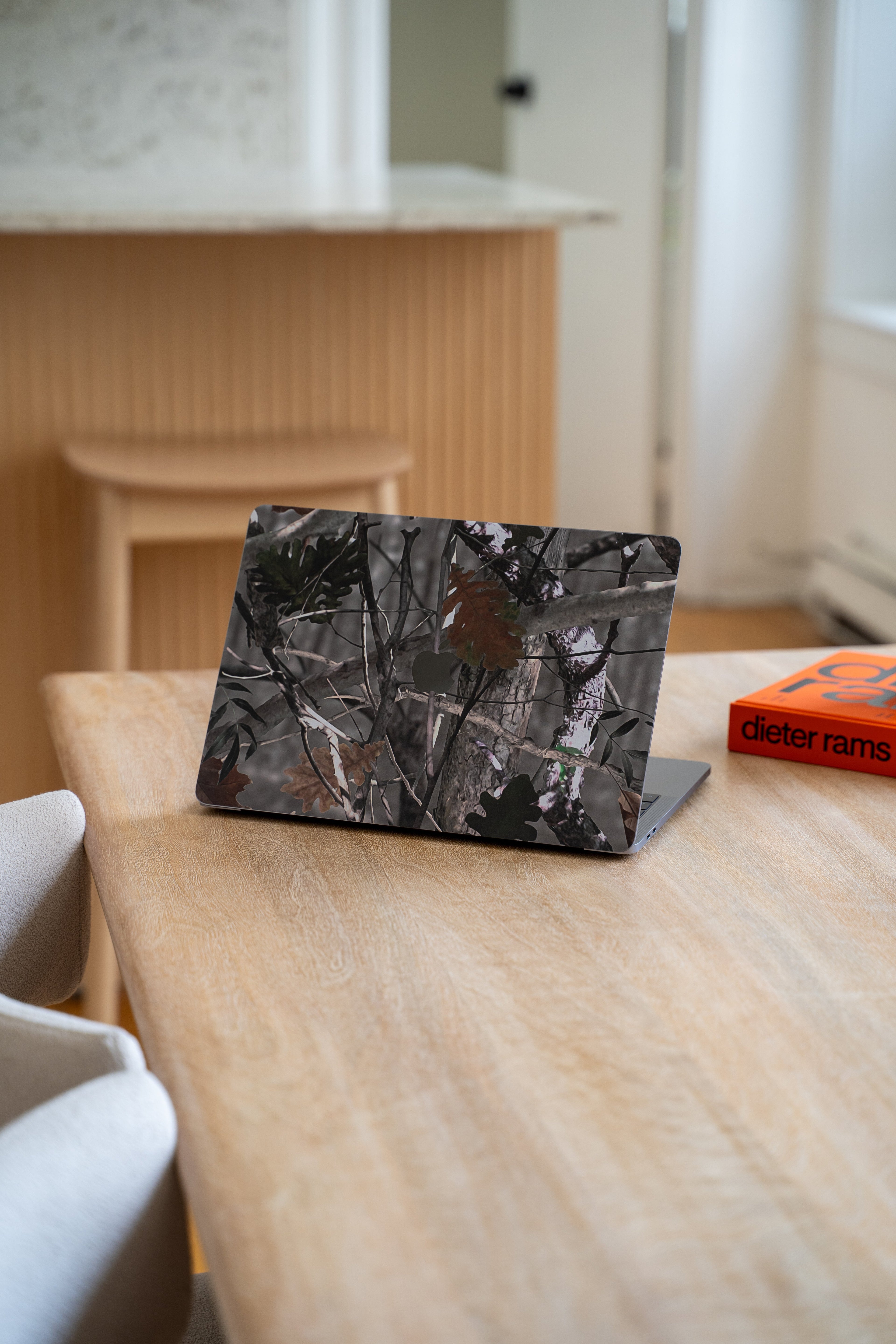 Laptop with a forest camouflage skin sitting on a light wood table in a modern, minimalist interior.