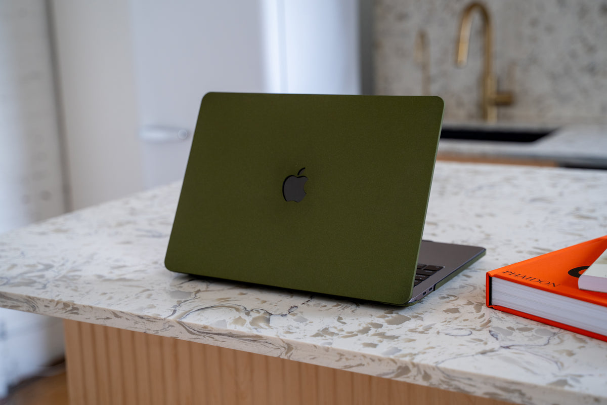 Olive green matte laptop skin on a MacBook resting on a marble kitchen island next to an orange book.