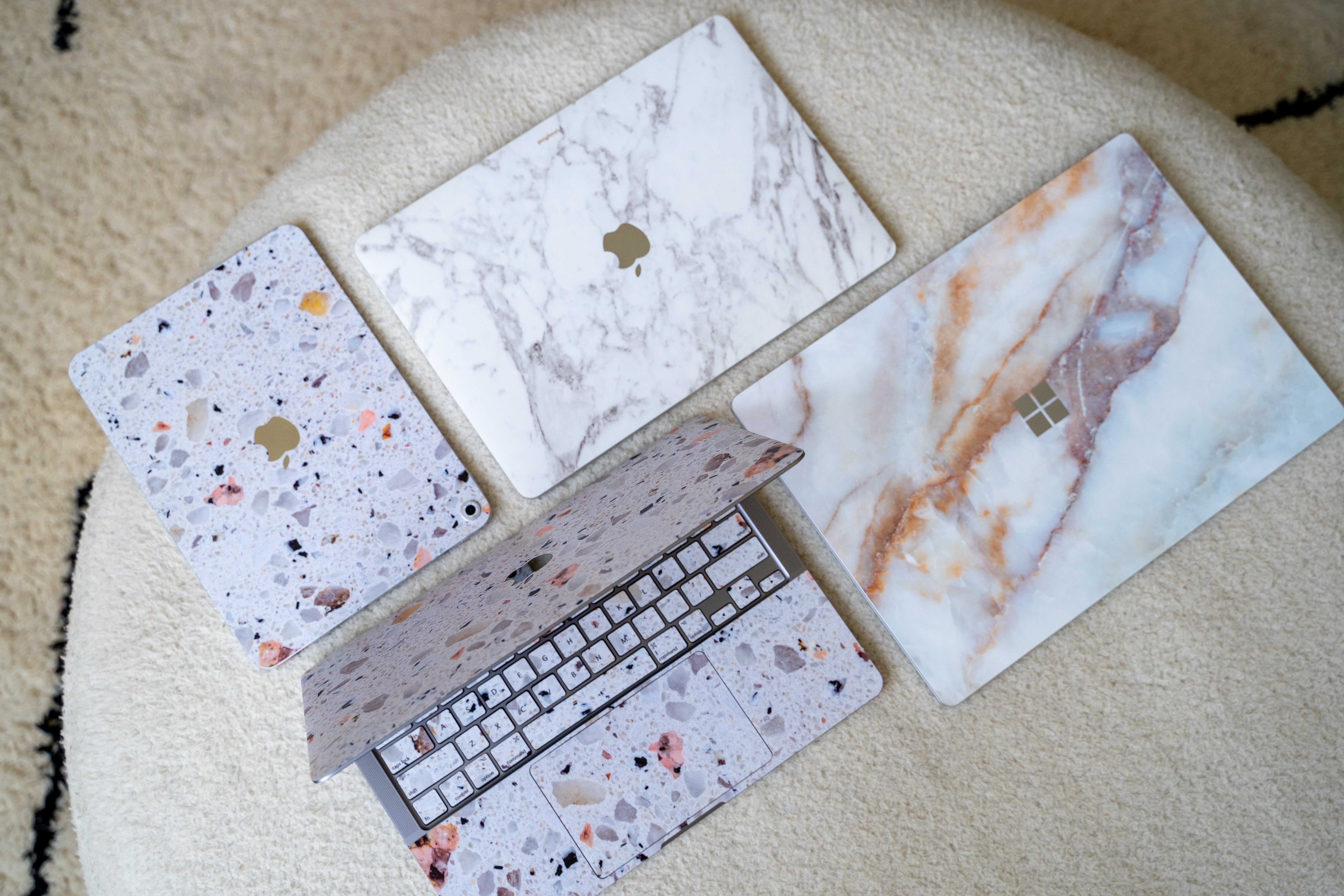 Laptops and tablets featuring decorative marble and terrazzo skins arranged on a textured cream ottoman.