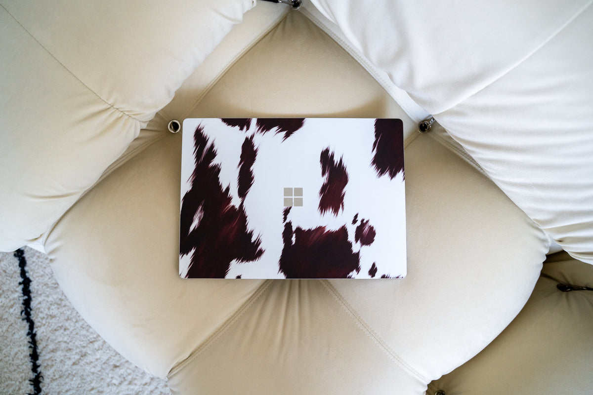 Microsoft laptop with a brown and white cow print skin resting on a cream-colored tufted leather chair.