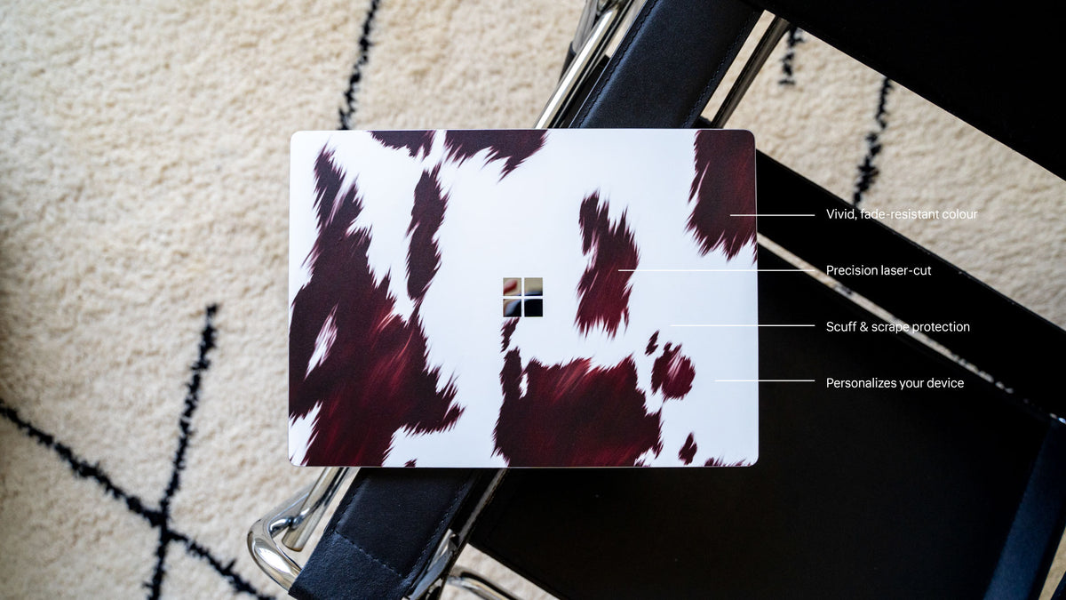 Laptop skin with a dark brown and white cow print pattern, featuring a precision laser-cut Microsoft logo cutout.